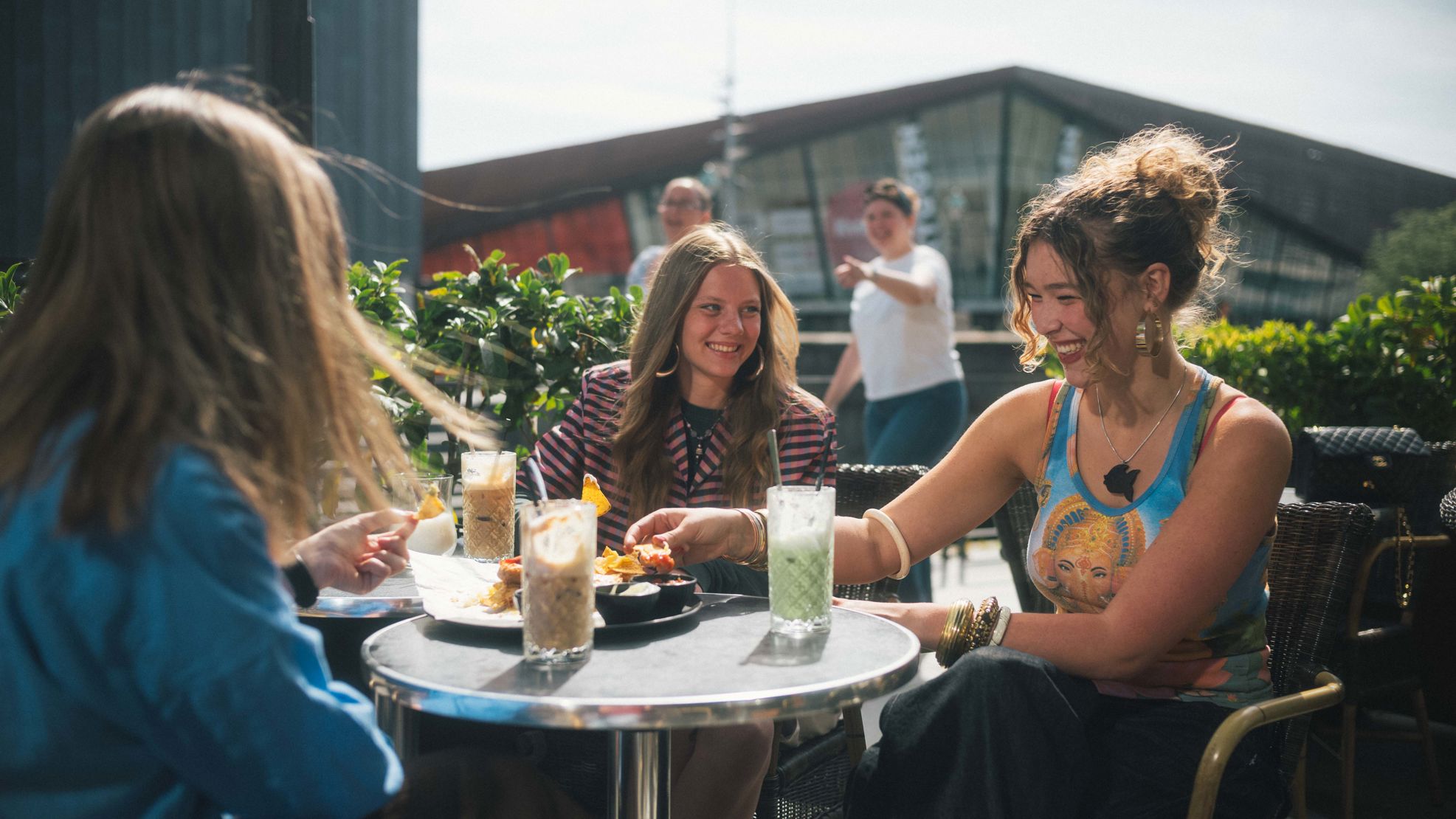 Tre unge kvinder sidder udenfor en café og snakker. Der er med og drikke på bordene imellem dem.